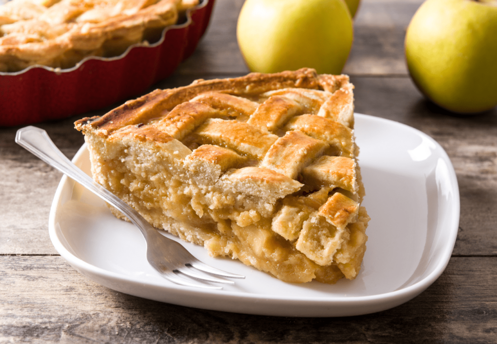 Slice of apple pie on a plate with a fork beside apples